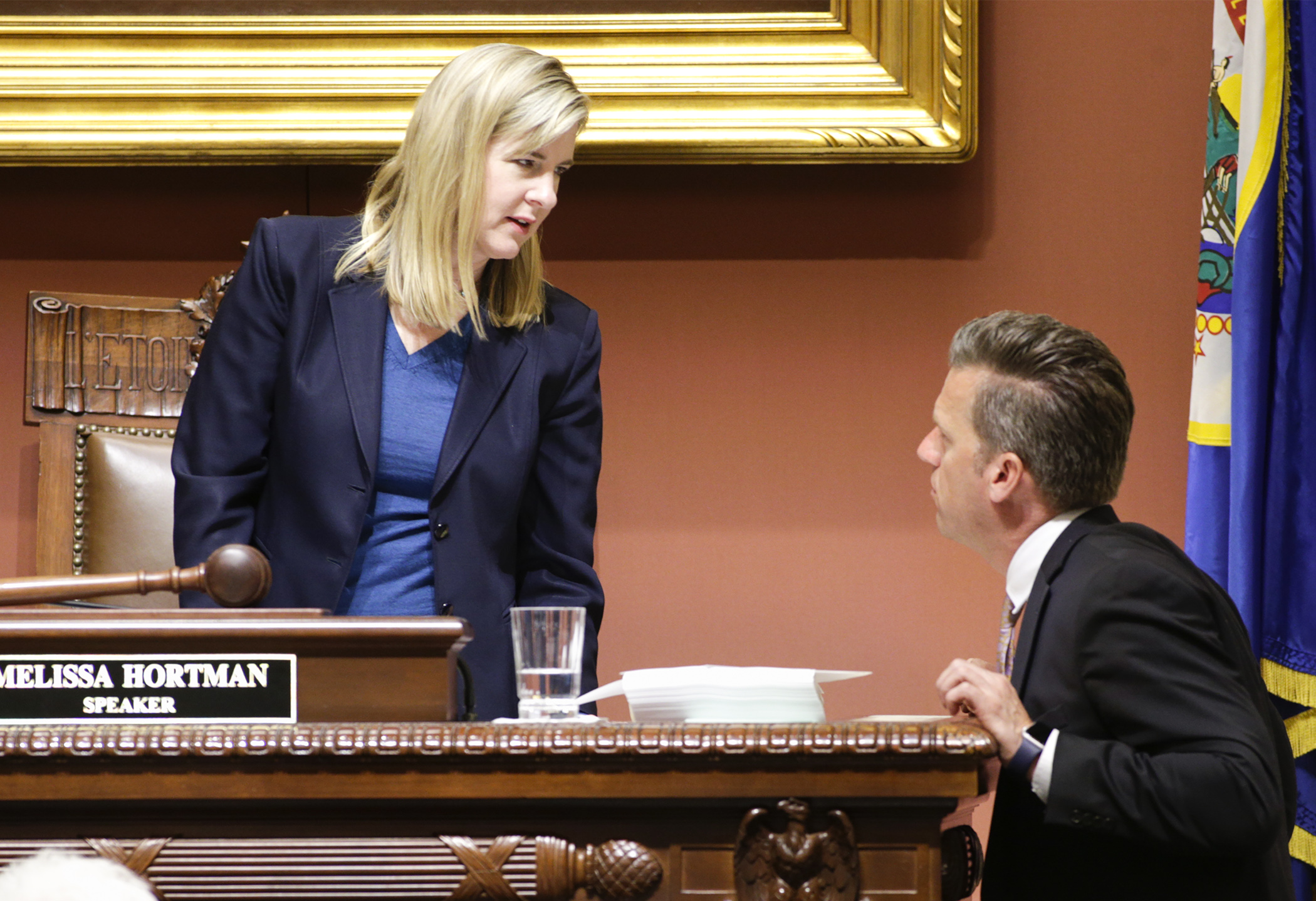 Speaker Melissa Hortman stands behind the Speaker's podium and speaks to Minority Leader Kurt Daudt, who stands beside the podium.