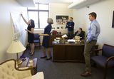 Representative Klevorn looks at a map that Claire Eickholt hangs up on a wall. Three men stand behind them, also looking at the map.
