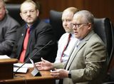 Scott Carlson sits at the testifiers' table and speaks to someone out of view. Four men sit next to him, one of whom is Representative Brand, who watches him as he speaks.