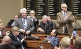 Representative Rarick smiles and stands at his House desk wearing a "powdered wig." Representative Nash stands at his House desk, applauds, and looks at someone out of view. Members sit around the two men, applauding and taking photos with their cell phones.