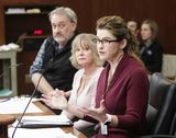 Kristy Beaucage sits at the testifiers' table and speaks to someone out of view while gesturing with her hands. A woman and Representative Hansen sit next to her and watch as she speaks.