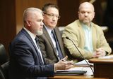 Chris Lennox sits at the testifiers' table and speaks to someone out of view. A man and Representative Freiberg sit next to him.