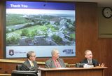 David Kelliher sits at the testifiers' table and speaks to someone out of view. Representative Urdahl and Kent Whitworth sit beside him and watch as he speaks. Behind all of them is a presentation screen that says, "Thank you."