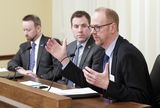 A man sits at the testifiers' table and speaks to someone out of view. Representative Long and another man sit next to him.