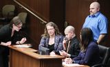 A young person, sitting at a table with two others, testifies using sign language. A sign language interpreter stands to the left, speaking into a microphone.