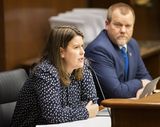 Jessica Webster sits at the testifiers' table and speaks to someone out of view. Representative Jurgens sits next to her and watches as she speaks.