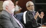 Theartrice Williams sits at the testifiers' table and speaks to someone out of view while gesturing with his hands. Representative Considine sits next to him.