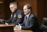 Representative Marquart sits at the testifiers' table and speaks to someone out of view. A uniformed officer sits beside him.