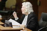 Representative Murphy sits at the testifiers' table and speaks to someone out of view while gesturing with her hand.