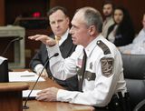 Tim Leslie sits at the testifiers' table and speaks to someone out of view while gesturing with one hand. Representative Lesch sits next to him and watches as he speaks.