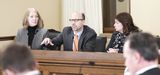 Representative Pinto sits between two people at the testifiers' table and speaks to someone out of view while gesturing with one hand.