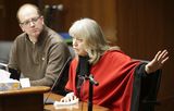 Mary Falk sits at the testifiers' table and speaks to someone out of view while gesturing with one hand. Representative Tabke sits next to her and watches as she speaks.