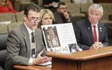 Tom Goeltz sits at the testifiers' table and speaks to someone out of view. Wendy Goeltz sits next to him and holds a photo of their daughter on a poster board. Representative Dettmer sits next to them and watches Tom as he speaks.