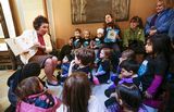 Representative Kunesh-Podein sits on a chair and holds a book open to the group of children sitting in front of her. The children are watching her read. Three adults sit and stand near the back of the group and also watch.