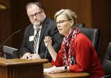 Representative Sandstede sits at the testifiers' table and speaks into a microphone to someone out of view. A person sits next to her and watches as she speaks.