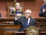 Governor Walz stands behind a podium in the House Chamber and speaks to someone out of view while gesturing with one hand.