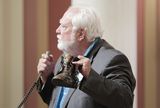 Representative Considine holds a microphone while speaking to someone out of view. In his other hand, he holds a man's boot.