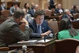 Sen. Aric Putnam sits at his Senate Chamber desk to have a conversation with two others, their backs to the camera.