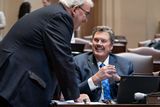 Senator Weber sits at his desk, smiling while looking at Senator Utke, who is slightly leaning over the desk.